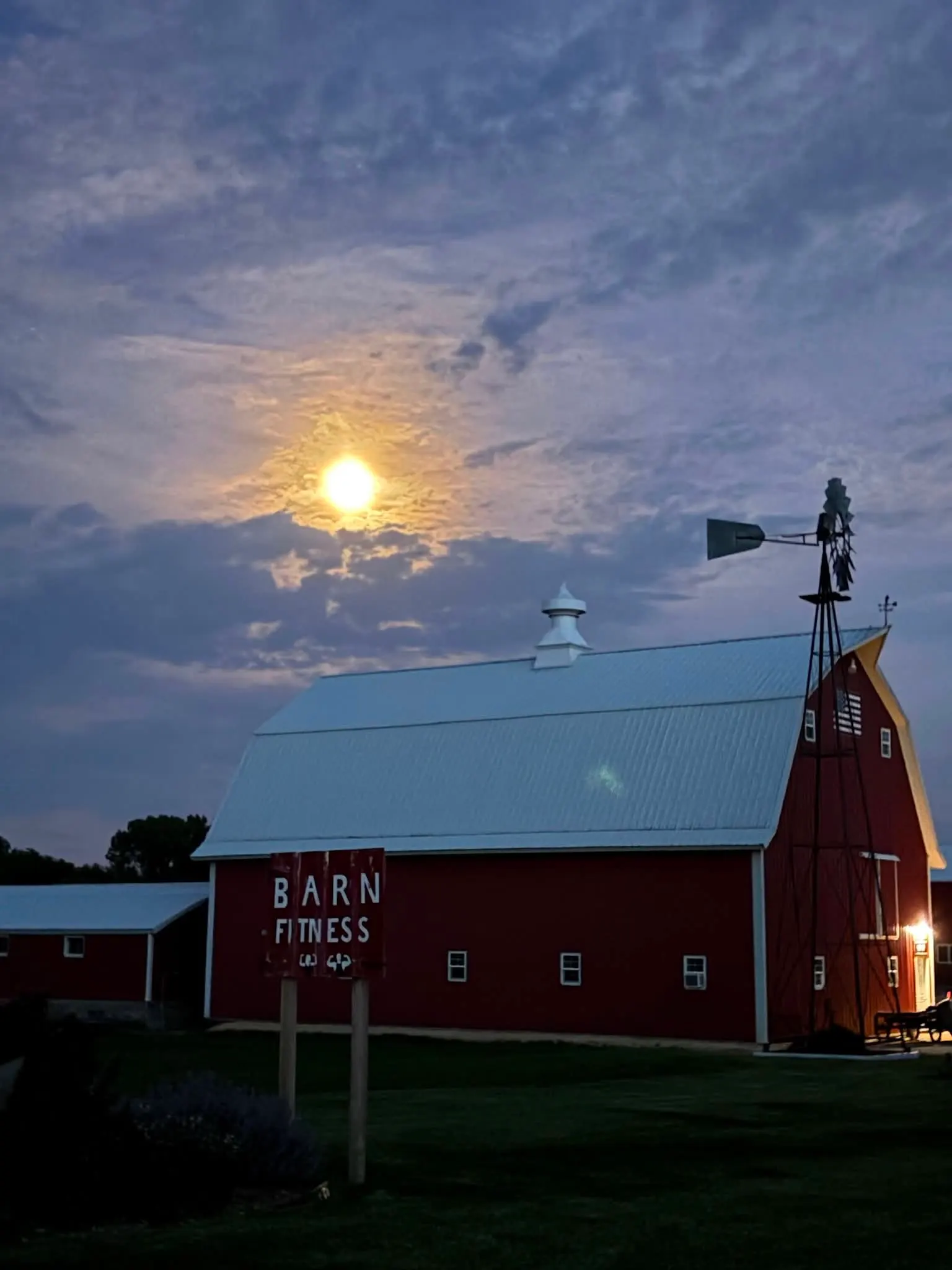 The red Barn Fitness barn and windmill at dusk under a full moon, with the Barn Fitness sign out front